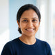 Smiling South Asian woman with dark hair, looking at the camera, against a soft blue and white background.