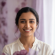 A smiling South Asian woman in her late 20s, looking confidently at the viewer, with a soft, blurred background in dusty lila